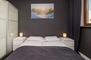 A bedroom with a bed, two bedside tables with lamps, a painting of a sunset over water, and a window with curtains.