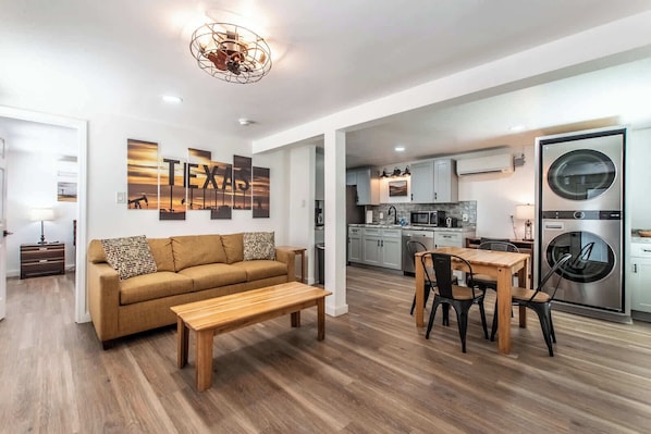 A modern living room with a sofa, coffee table, and dining area, connected to a kitchen with appliances and a laundry machine.