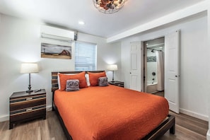 A bedroom with a large bed, two bedside tables, a nightstand, a ceiling light fixture, a window with blinds, and a door leading to a bathroom.