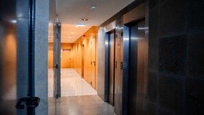 A modern hallway with a glass door on the left, leading to a room with a wooden door. There are two elevators on the right, and the floor is tiled. - Stunning balcony with views of Tagus River, ocean and historic sites of Belém✨ (Lisboa)