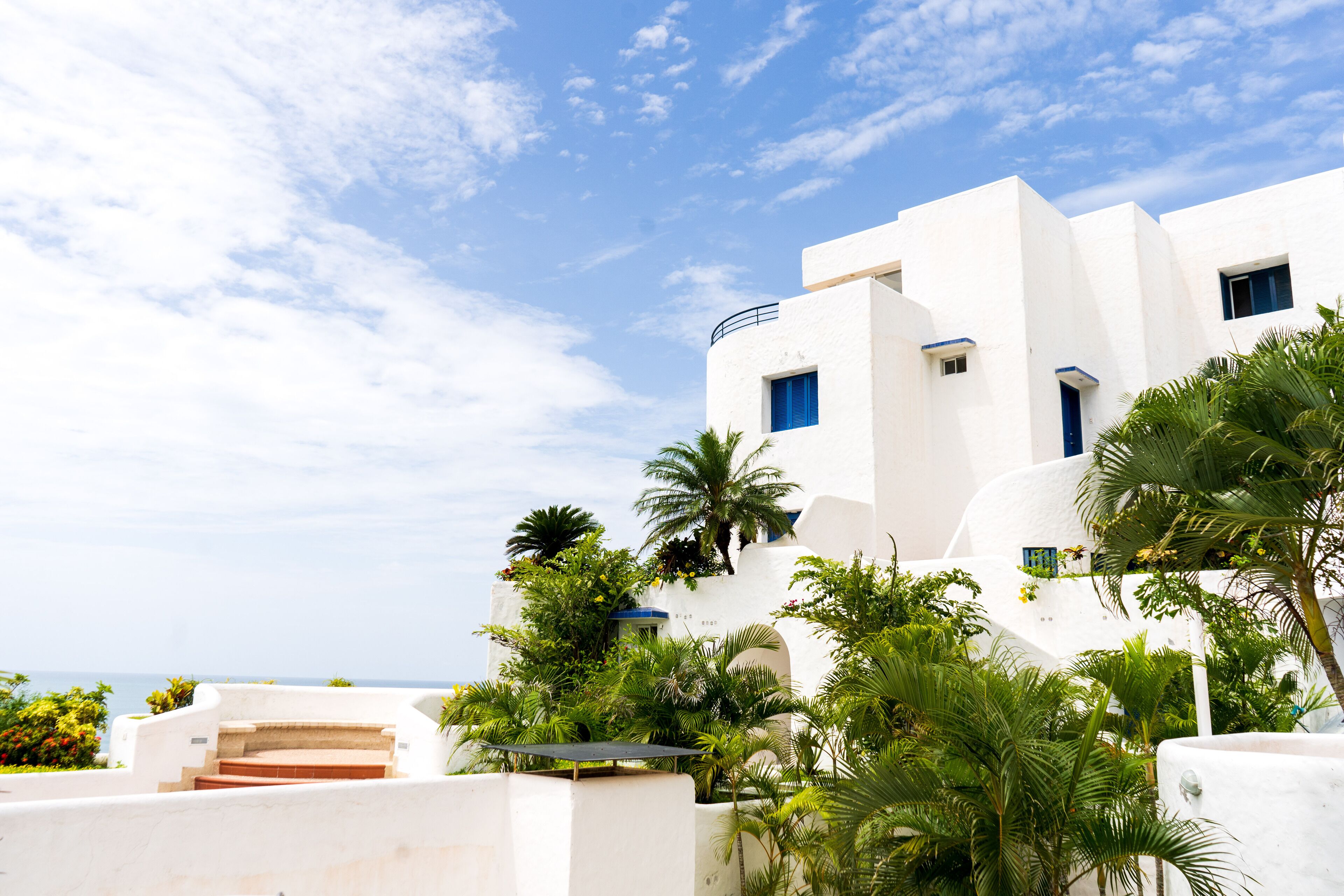 A white building with a balcony, surrounded by palm trees and a clear sky.