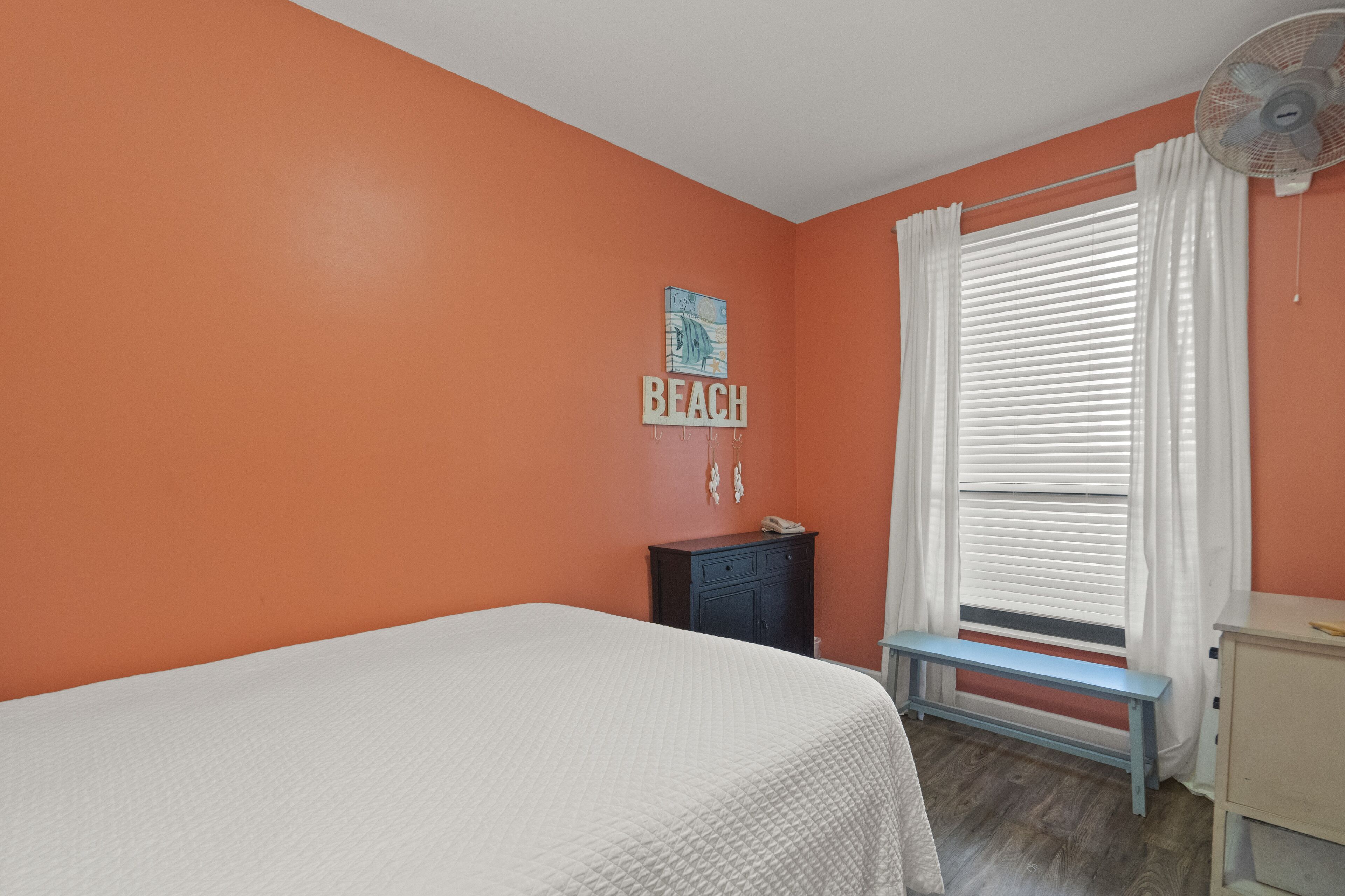A bedroom with an orange wall, a bed, a nightstand, a bench, and a window with white curtains.
