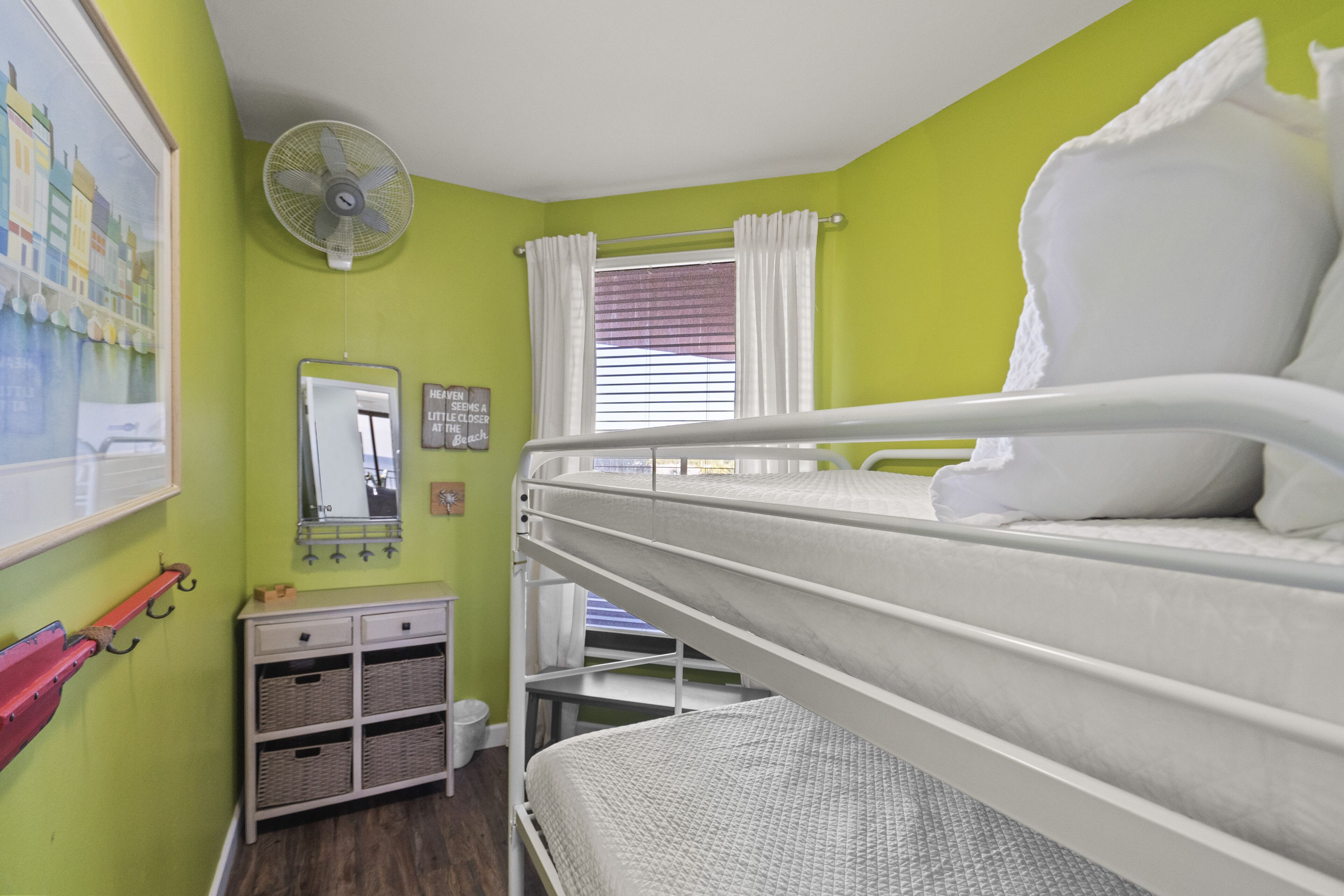 A bunk bed room with a fan, a window with curtains, a mirror, and a small cabinet with baskets.