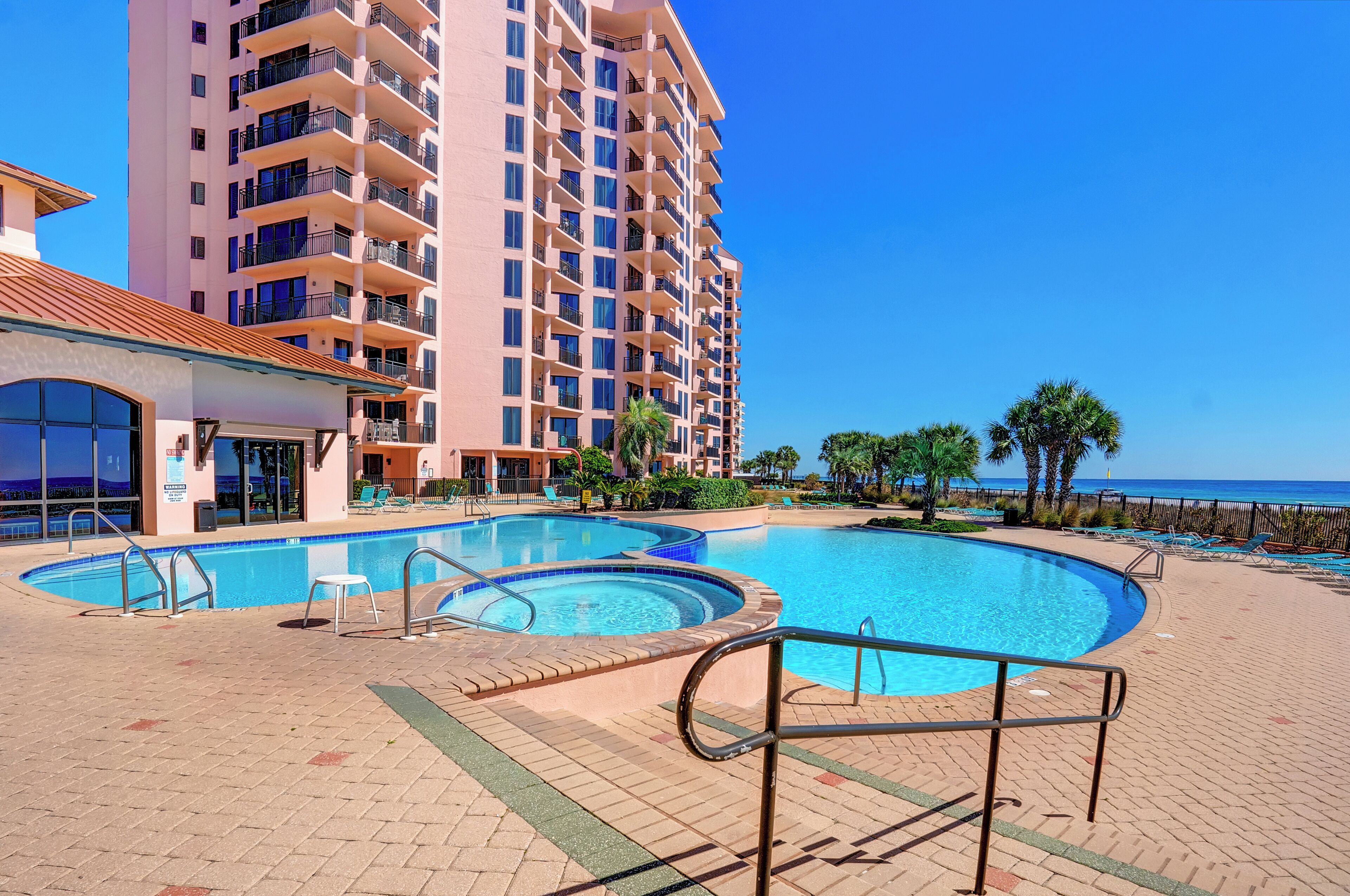A hotel with a large swimming pool, a beach view, and a tall building in the background.