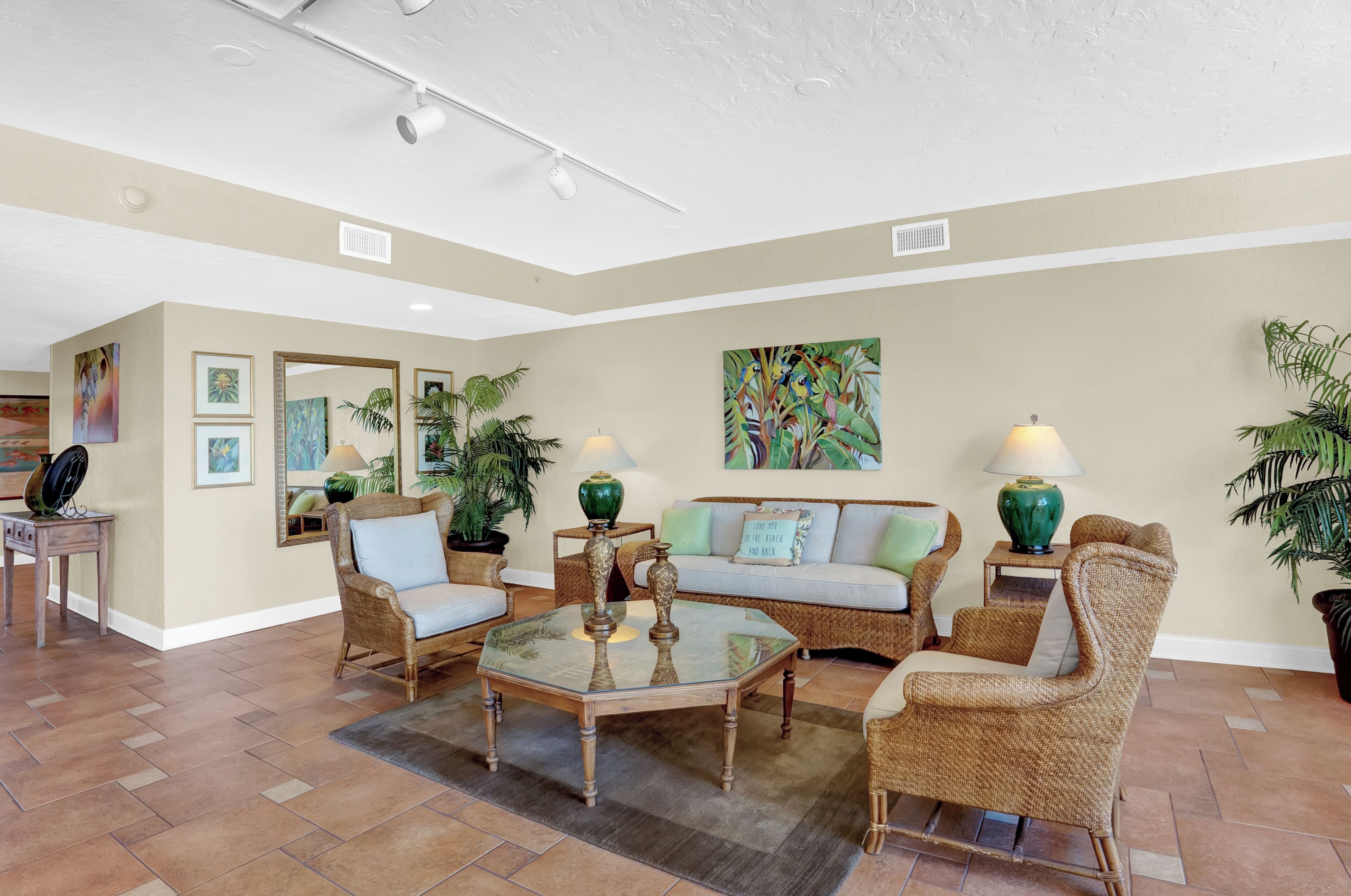 A spacious living room with a glass coffee table, wicker furniture, and several potted plants.