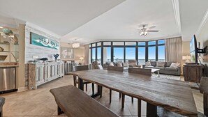 A spacious living area with a large wooden dining table, multiple sofas, and a view of the outdoors through large windows. - Beachfront!! Lazy River and Slide!!  New Rental!! (Orange Beach)