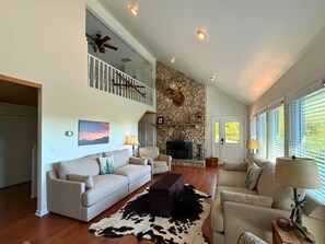 A spacious living room with a fireplace, a staircase, and a large window with blinds.