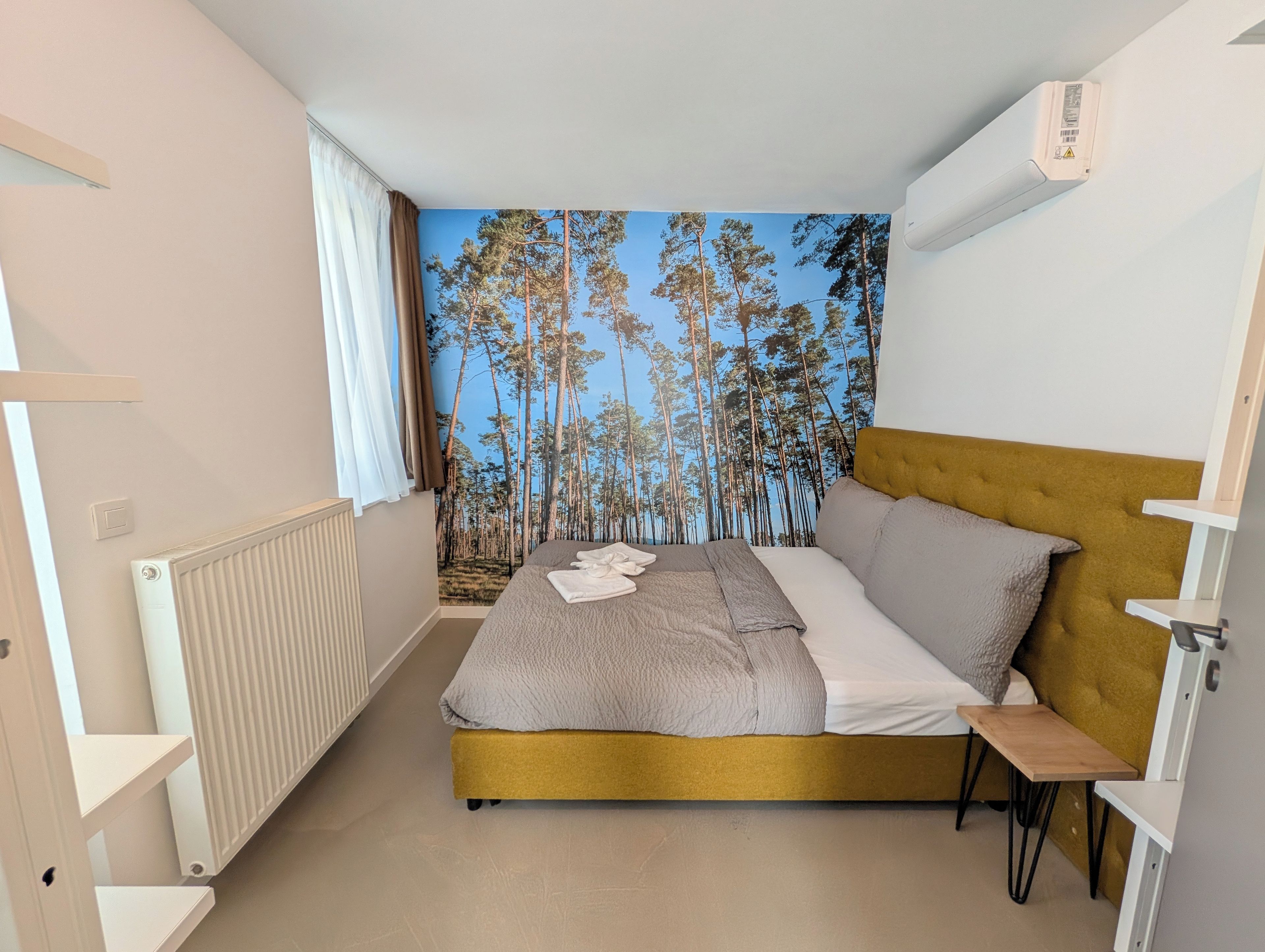A bedroom with a bed, a radiator, a window with curtains, and a wall with a forest mural.