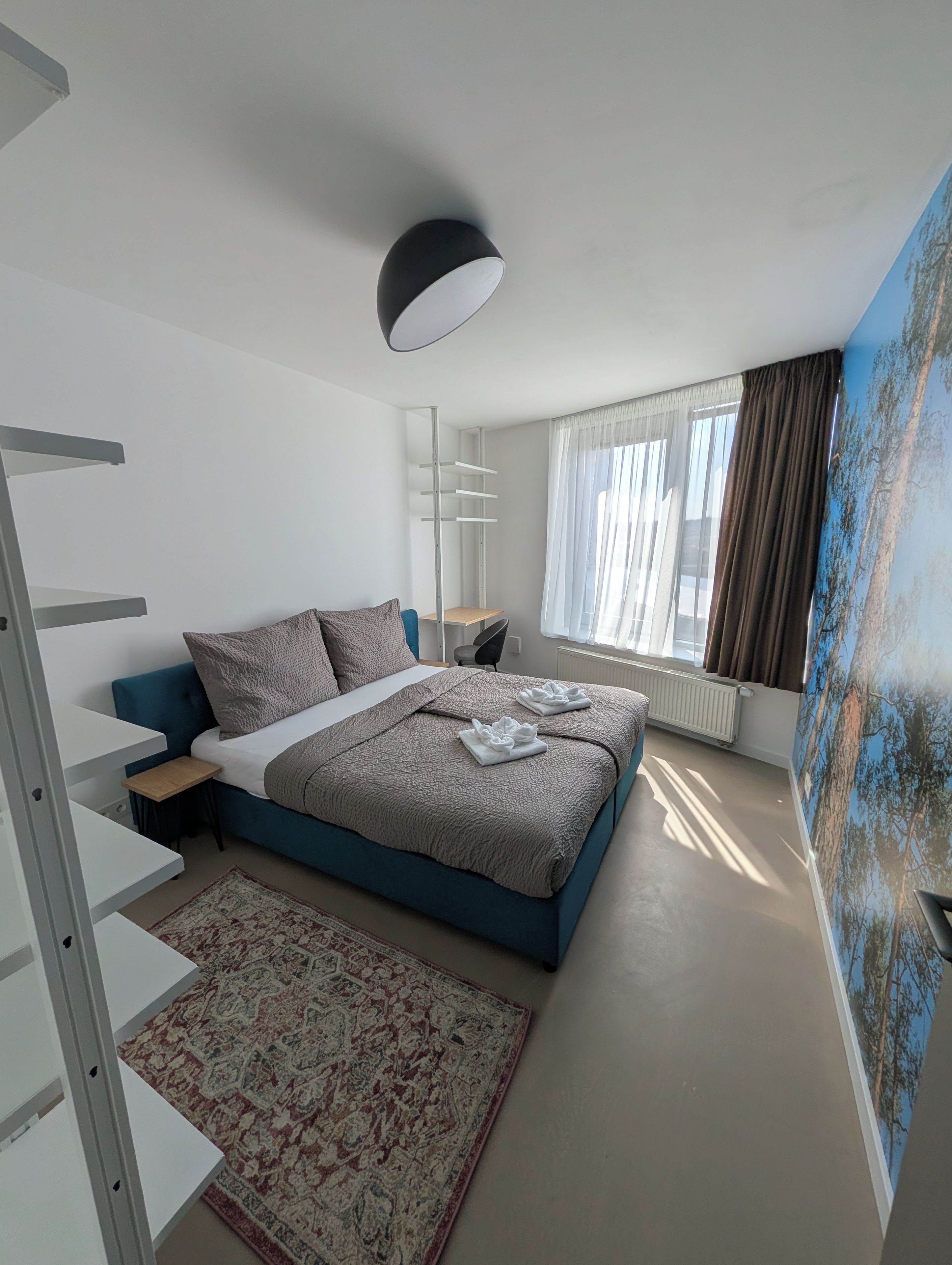 A modern bedroom with a bed, a desk, a chair, a radiator, a window with curtains, and a painting on the wall.