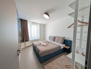 Un dormitorio con una cama, una mesita de noche, un radiador, una ventana con cortinas y una lámpara en el techo.