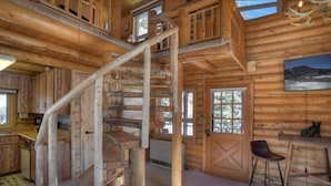 L’intérieur d’une cabane en rondins, avec un escalier en bois, un coin repas et une grande fenêtre donnant sur un paysage magnifique.