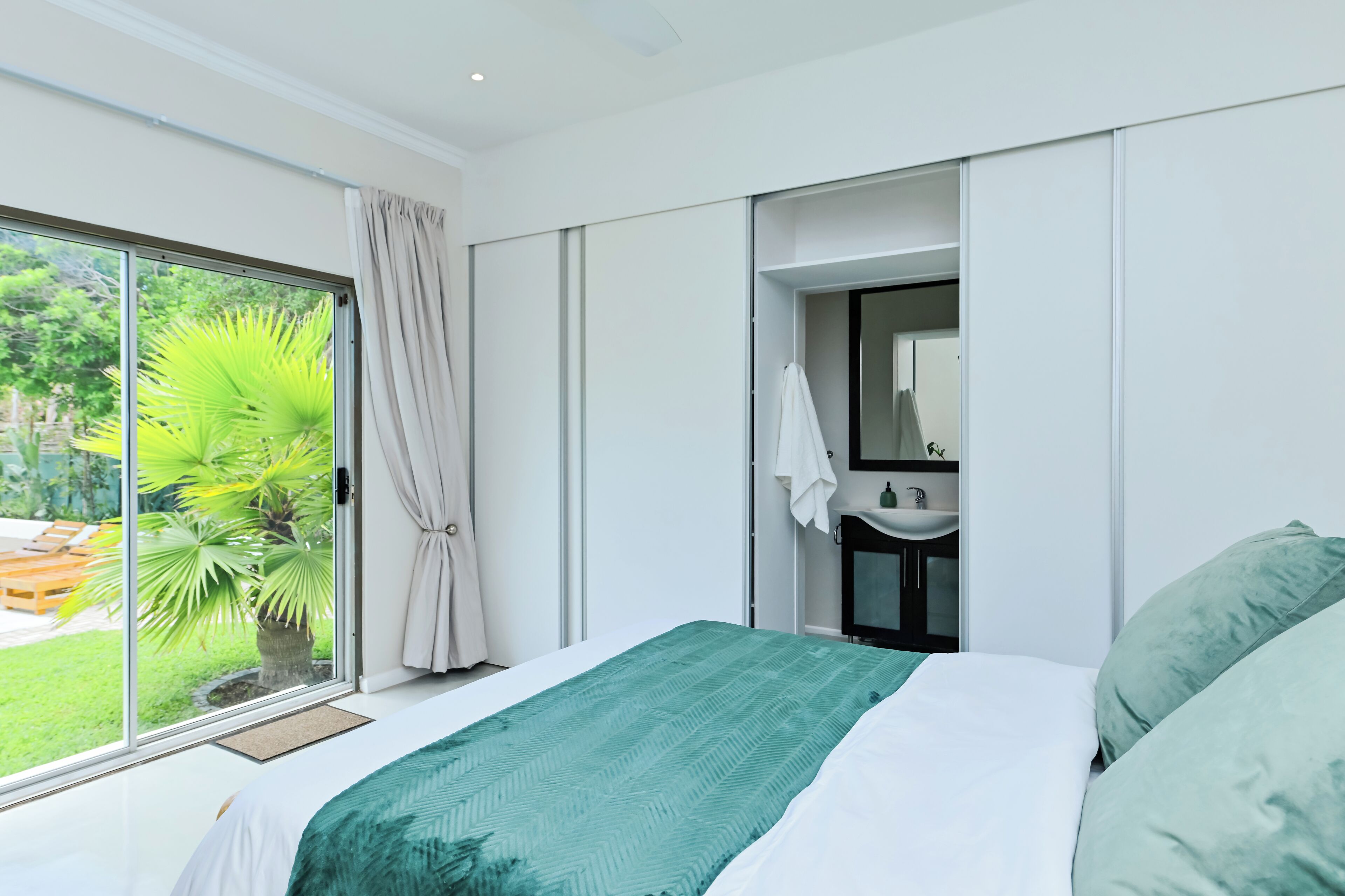 A modern bedroom with a large bed, a bathroom with a sink, and a view of greenery outside.