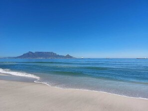 A beach with clear blue water, a sandy shore, and a distant mountain. - Spray Villa Bloubergstrand - with hot tub and pool, only 500m from the beach! (Bloubergstrand)