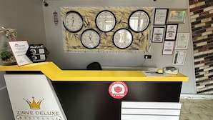 A reception desk with a yellow top, a black base, and a red circular logo. There are clocks on the wall behind the desk, and a red carpet with a gold pattern leads to the desk. - Zirve Suit Rezidans (Talas)