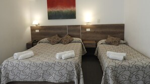 Habitación de hotel con dos camas, cada una con una colcha estampada, cabeceros, lámparas de noche y un cuadro en la pared.