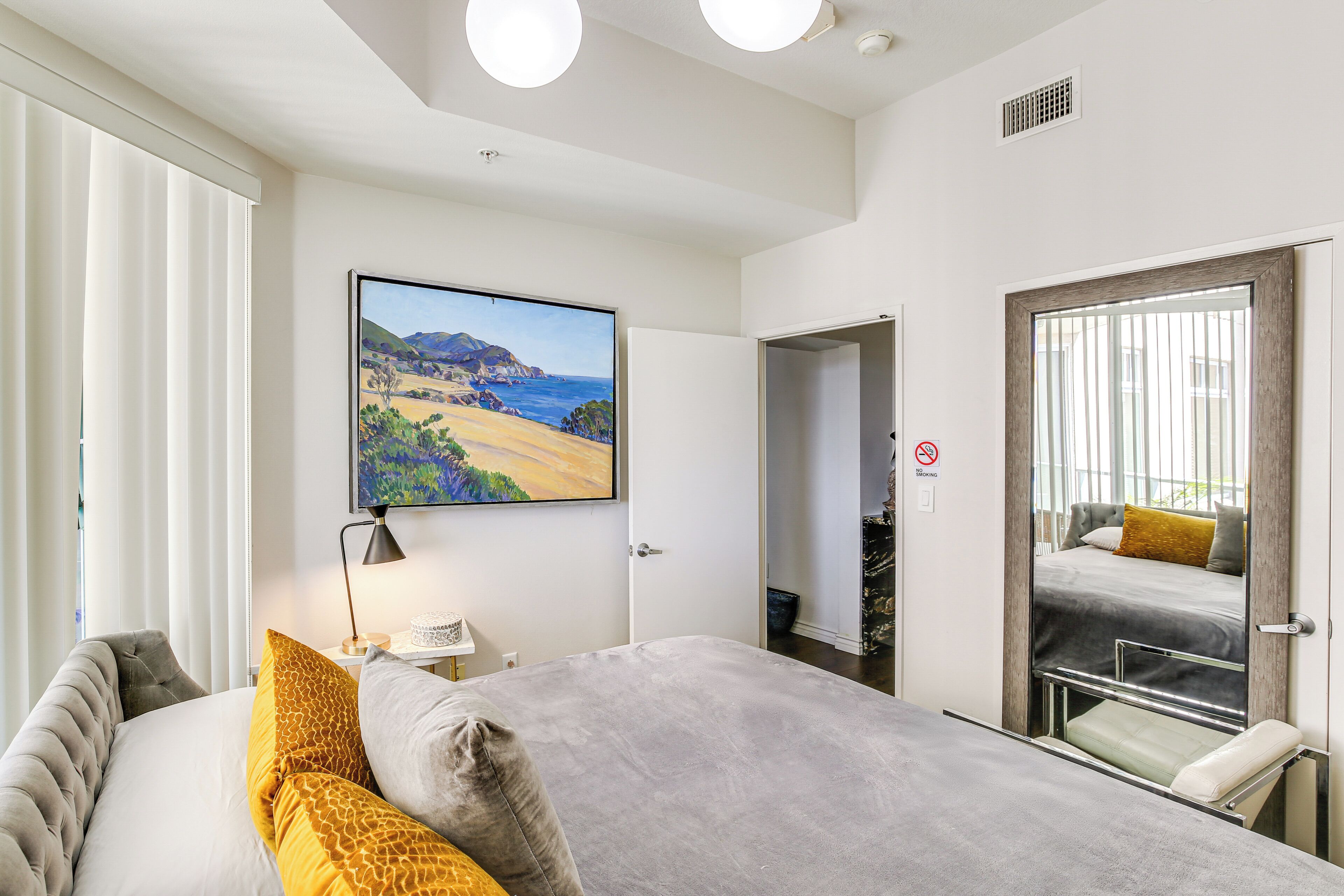 A modern hotel room with a large bed, a painting of a landscape, a desk with a lamp, and a view of the outdoors through a door.