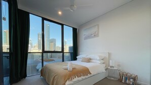 A modern hotel room with a large bed, a view of the city skyline through floor-to-ceiling windows, a ceiling fan, and a bedside table with a lamp and a magazine rack. - Charming Urban Retreat (South Brisbane)