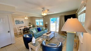 A living room with a sofa, coffee table, and chairs. There is a dining area with a table and chairs. A ceiling fan and a lamp are visible. - Beach Access, Lagoon, Fishing Pier and Pool (Gulf Shores)