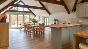 Een ruime keuken met houten balken, een eethoek met tafel en stoelen, en uitzicht op de omgeving door grote ramen.
