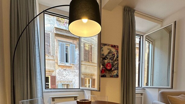 A dining area with a round table, clear chairs, a modern floor lamp, and a view of a building with shutters through large windows. - Newly renovated, located on the 2nd floor in a very quiet area of Trastevere. (Roma)