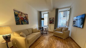 A living room with a beige sofa, a smaller armchair, a coffee table, and a lamp. There are two windows with curtains, a painting on the wall, and a television. - Newly renovated, located on the 2nd floor in a very quiet area of Trastevere. (Roma)