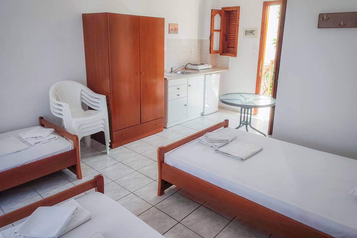 A hotel room with two beds, a wardrobe, a small kitchenette, a chair, and a table.
