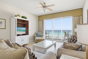 A living room with a television, a ceiling fan, a sofa, a coffee table, and a view of the ocean through large windows.