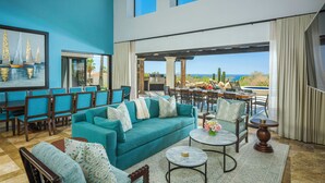 Un vaste espace de vie agrémenté d’un canapé turquoise, de fauteuils assortis et offrant une vue sur l’océan grâce à de grandes fenêtres.