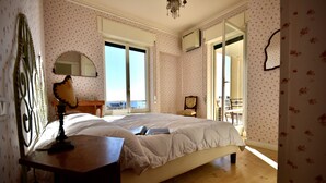 A bedroom with a large bed, a wooden nightstand, a mirror, and a window with a view. - Panorama Apt in Ospedaletti by Wonderful Italy (Ospedaletti)