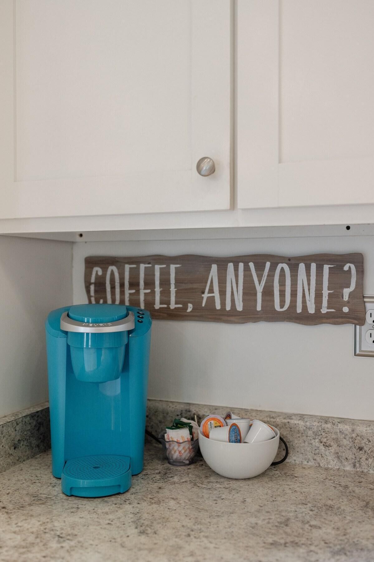 Una cocina con cafetera, taza y un letrero que dice 'COFFEE, ANYONE?'.