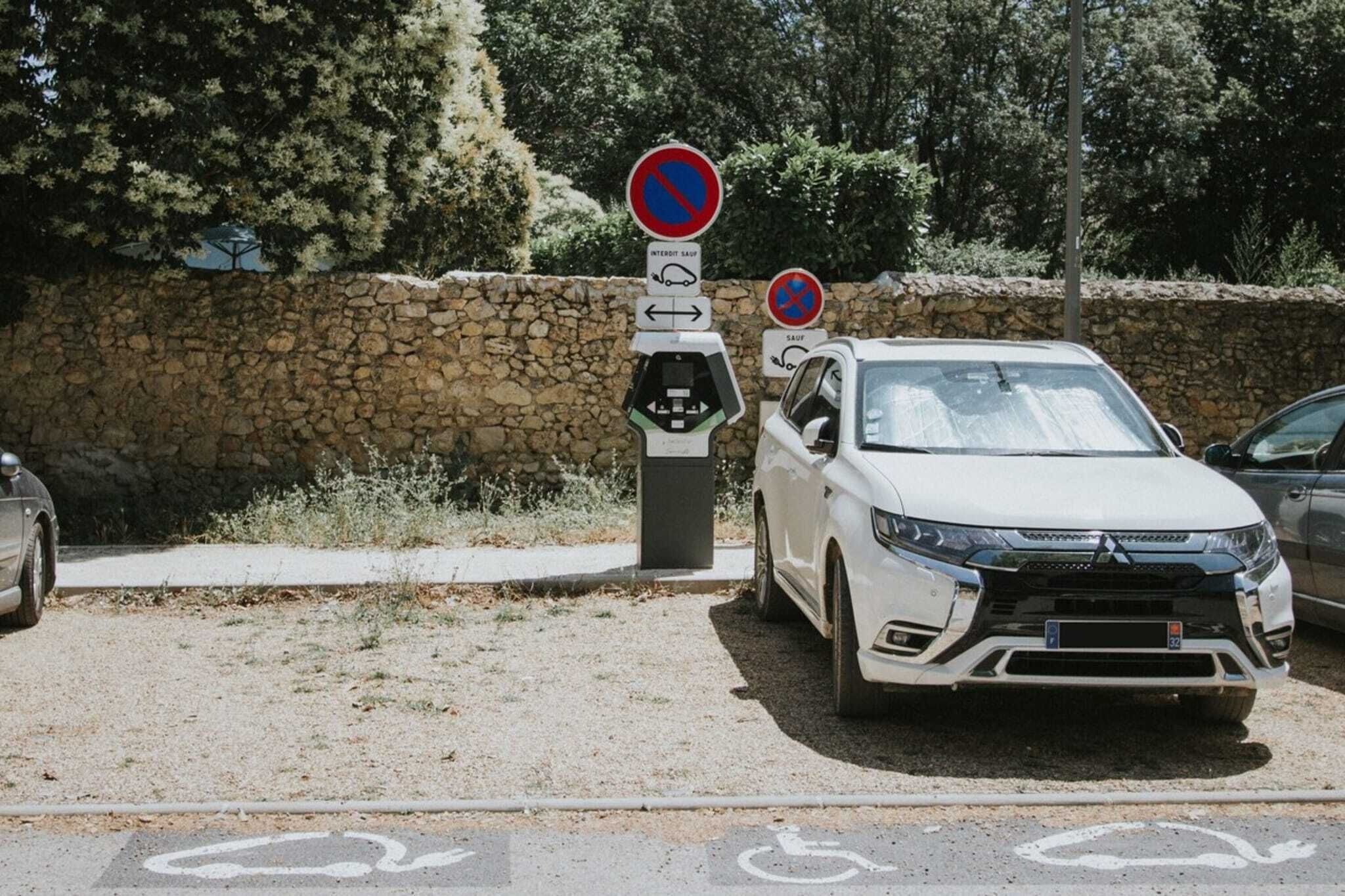 Un coche blanco estacionado en un peaje, con un muro de piedra y árboles al fondo.