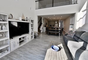 Ein modernes Wohnzimmer mit Flachbildfernseher, einer Holzwand zur Unterhaltungselektronik, einem grauen Sofa und einem Essbereich mit Treppe.