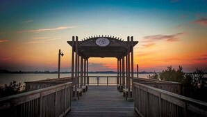 Een houten steiger die leidt naar een prieel met een cirkelvormig embleem, met uitzicht op een wateroppervlak bij zonsondergang.