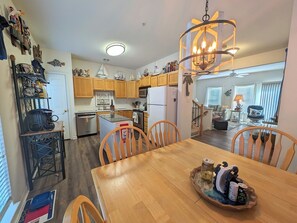 Cozinha com armários de madeira, uma mesa de jantar com cadeiras, um lustre e vista para a sala de estar.