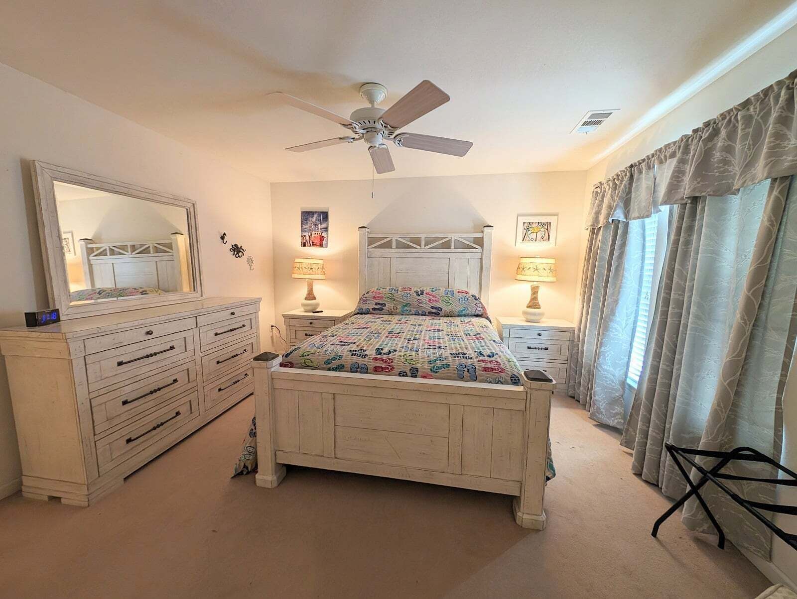 A bedroom with a bed, dresser, mirror, ceiling fan, two bedside tables with lamps, and curtains.
