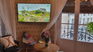 A room with a TV on the wall, a patterned chair, a small table with a vase of flowers, and a view of the outside balcony.