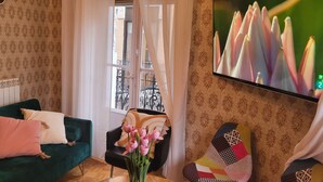 A living room with a green sofa, a black chair, a TV on the wall, and a window with a view of buildings.