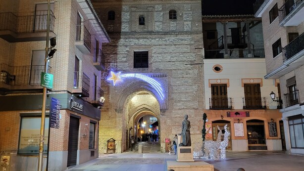 A historic street with stone buildings, a statue, and illuminated arches.