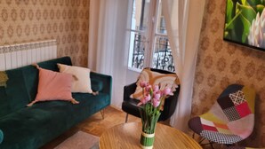 A living room with a green sofa, a small round table with a vase of flowers, a black chair, and a patterned armchair.