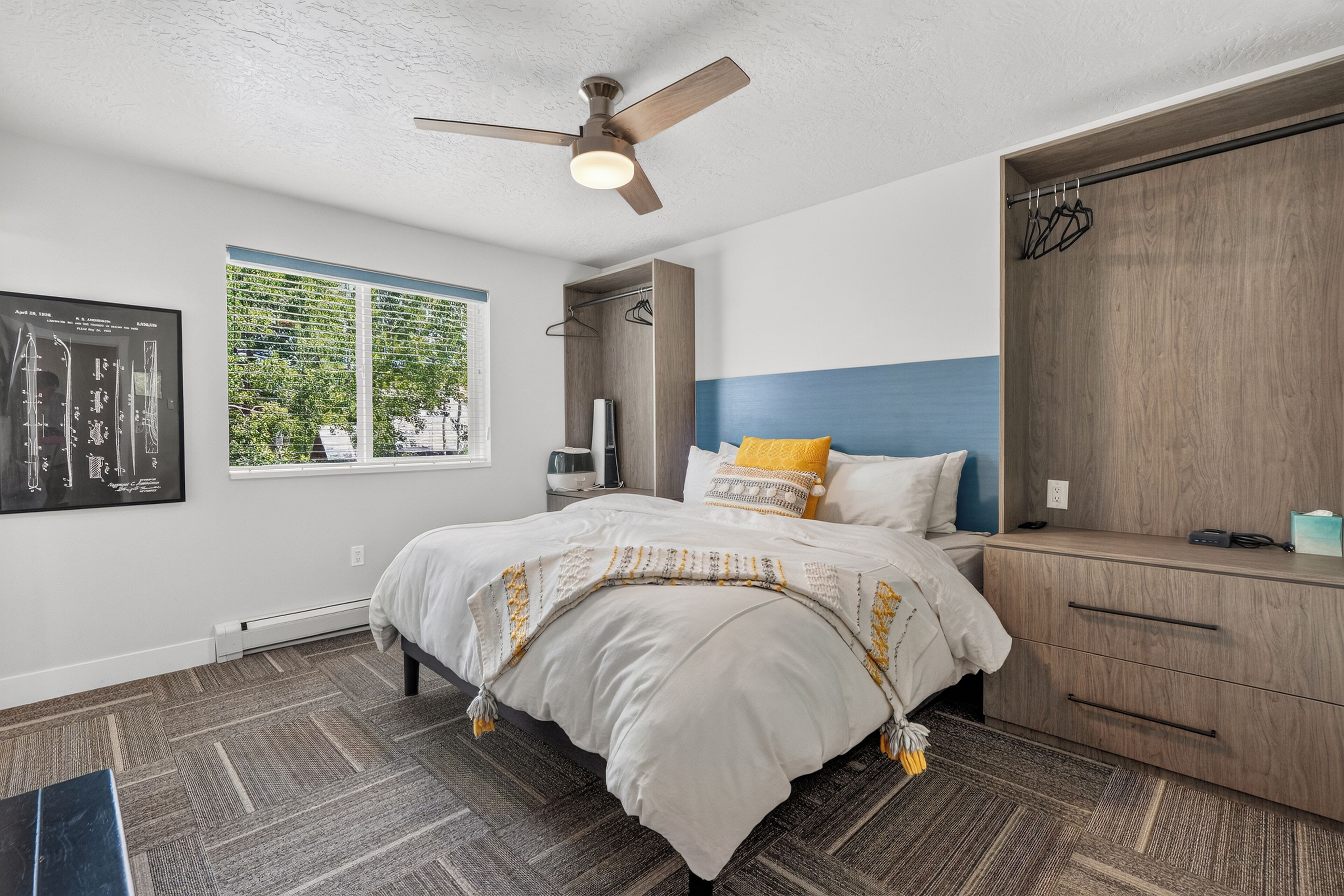 A bedroom with a bed, a wardrobe, a ceiling fan, a window with blinds, and a wall-mounted artwork.