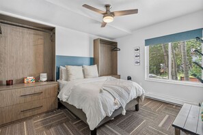 A bedroom with a bed, bedside table, wardrobe, ceiling fan, window with blinds, and a view of greenery outside.