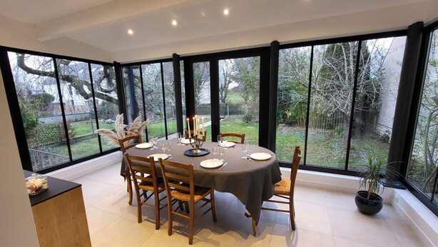 A dining area with a round table set for six, surrounded by large windows offering a view of the garden. - Createur DE Souvenirs (Chartres)