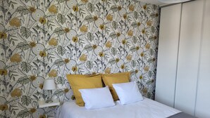 A bedroom with a floral wallpaper, a bed with white bedding and mustard pillows, a bedside table with a lamp, and a wardrobe. - Createur DE Souvenirs (Chartres)