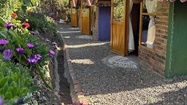 Una hilera de cabañas pequeñas y rústicas con puertas de madera y techos de metal rojo, rodeadas por un camino de grava y coloridos macizos de flores.