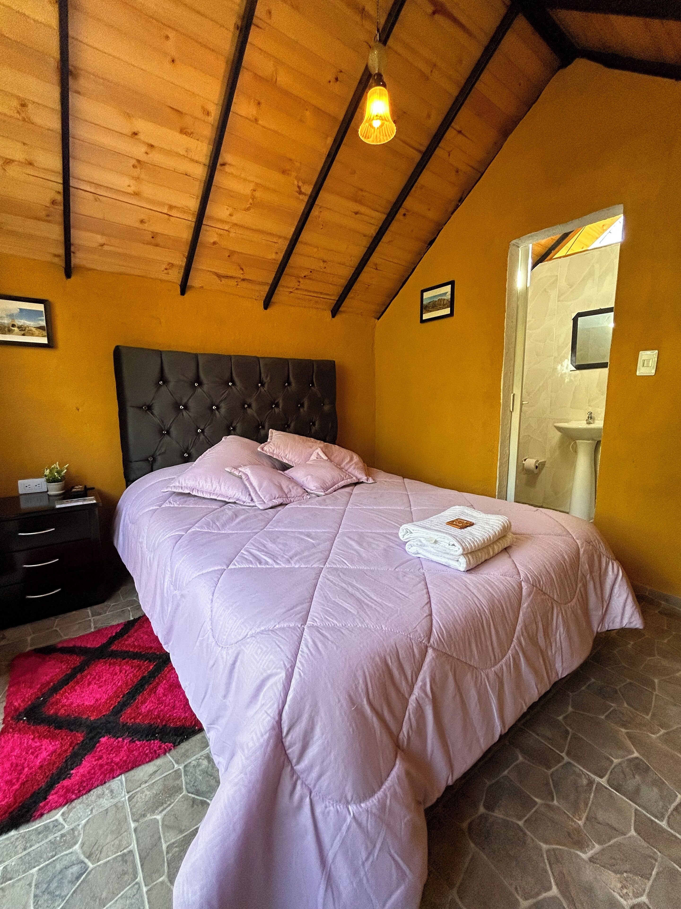 Een slaapkamer met een groot bed, een nachtkastje, een rood en zwart tapijt, een houten plafond, en een badkamer die door een open deur zichtbaar is.