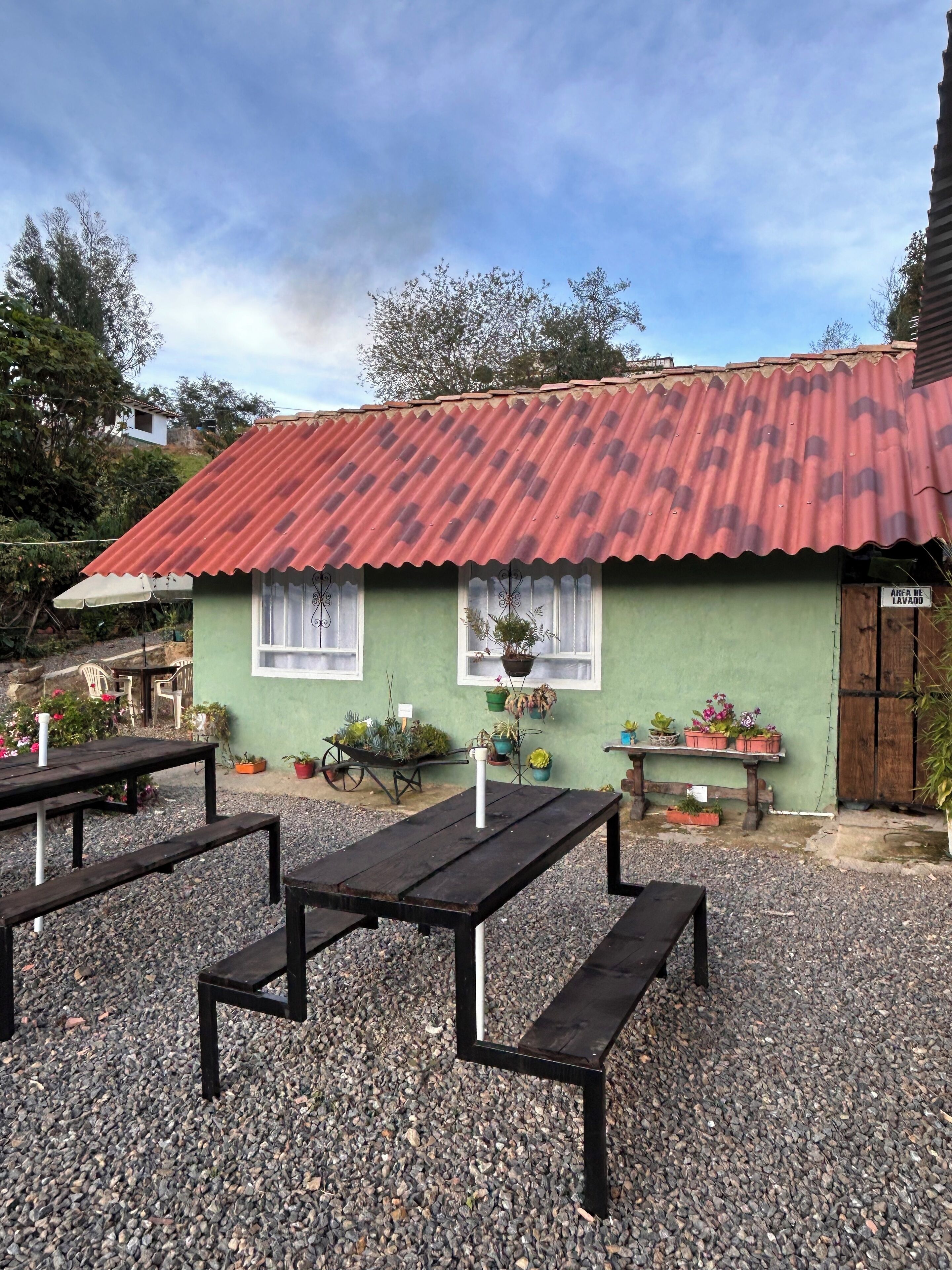 A small building with a red roof and green walls, surrounded by gravel and potted plants.