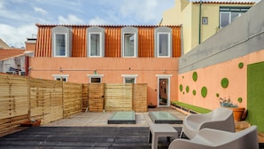 Un patio con valla de madera, una zona de estar pequeña y un edificio de paredes naranja y tejado de tejas.