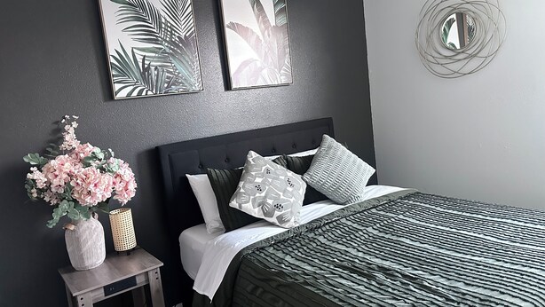A bedroom with a dark grey wall, a bed with a striped bedspread, multiple pillows, a small side table with a vase and a lamp, and two framed botanical prints on the wall. - Capital City- 6 Miles from Madison with fireplace (Sun Prairie)