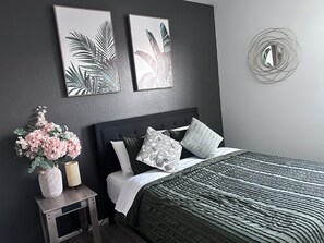 A bedroom with a dark grey wall, a bed with a striped bedspread, multiple pillows, a small side table with a vase and a lamp, and two framed botanical prints on the wall. - Capital City- 6 Miles from Madison with fireplace (Sun Prairie)