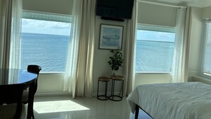 Habitación de hotel con cama, escritorio, dos sillas, televisor y ventanas amplias con vista al mar.